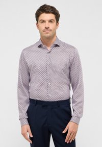Bluse mit Knöpfen, die ein geometrisches Muster in Rot und Marineblau auf einem hellen Hintergrund zeigt. Lange Ärmel, Kragen und taillierte Passform. Kombiniert mit dunklen Hosen.