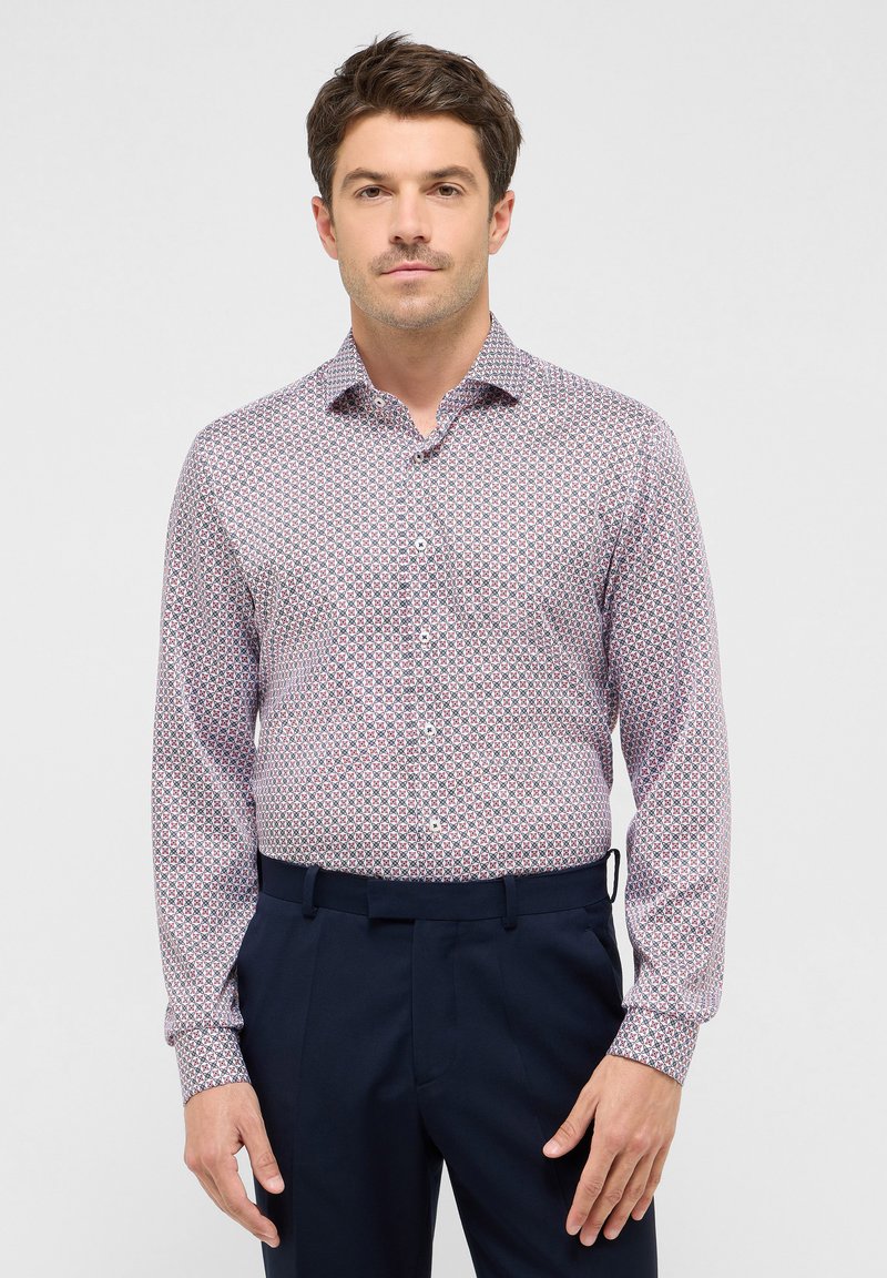 Bluse mit Knöpfen, die ein geometrisches Muster in Rot und Marineblau auf einem hellen Hintergrund zeigt. Lange Ärmel, Kragen und taillierte Passform. Kombiniert mit dunklen Hosen.