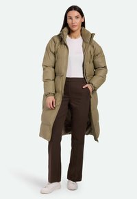 Abrigo acolchado largo de color verde oliva con cierre de cremallera, cuello alto y puños elásticos, combinado con pantalones marrones y zapatillas blancas.