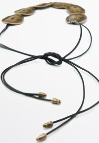 Dischi di metallo tono bronzo collegati da corde di pelle nera, con un nodo e tre estremità appuntite, che creano un design testurizzato e stratificato.
