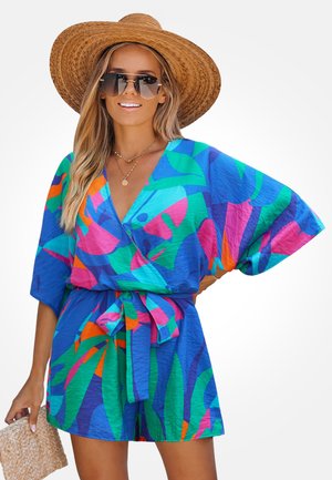 Frau trägt ein farbenfrohes Kleid mit blauem, grünem, pinkem und orangefarbenem Muster, weitem Ärmeln, einen breiten Strohhut, Sonnenbrille und hält eine gewebte Clutch.