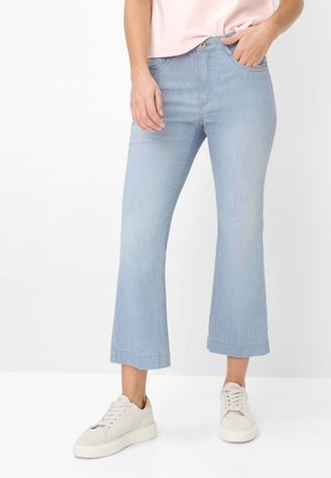 Person iført lyseblå cropped flare jeans, lyserød top og hvide sneakers, stående med den ene hånd i lommen.