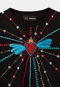 Czarna koszula z kolorowym haftem przedstawiającym serce z skrzydłami, kwiatowymi akcentami i geometrycznymi wzorami w czerwonych, niebieskich i złotych nitkach.