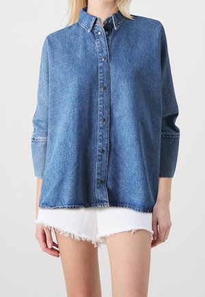 Camicia di jeans di blu medio, con chiusura frontale a bottoni, colletto e maniche lunghe. Orlo irregolare e dettagli di cucitura visibili.