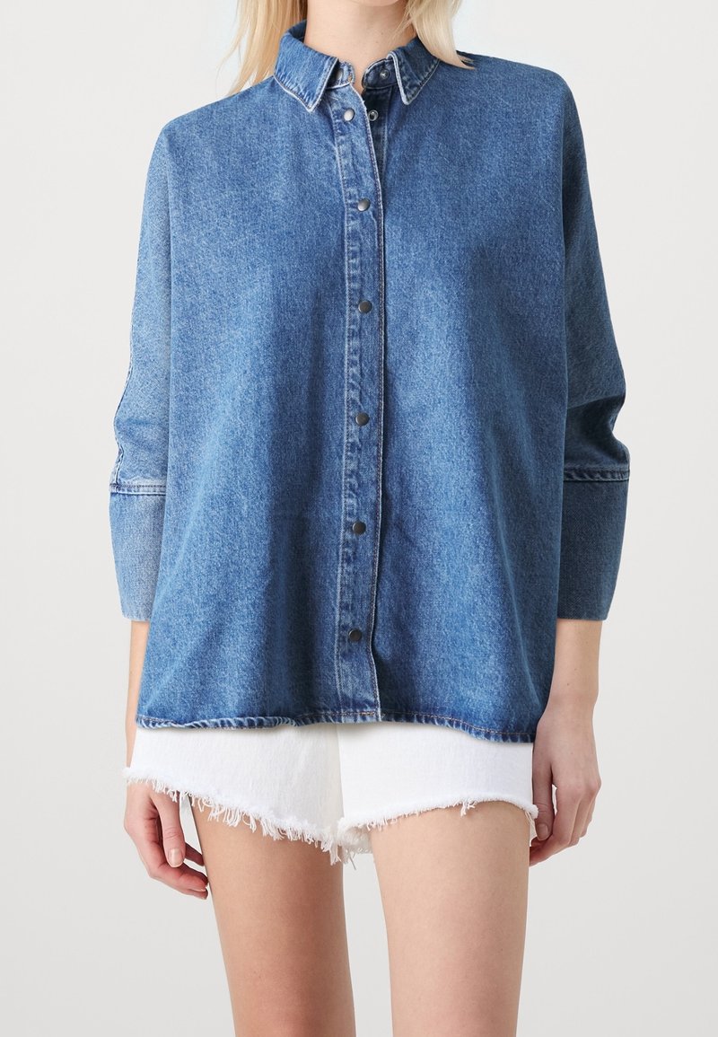Camicia di jeans di blu medio, con chiusura frontale a bottoni, colletto e maniche lunghe. Orlo irregolare e dettagli di cucitura visibili.