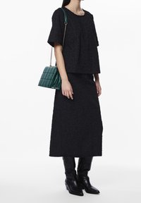 Woman wearing black textured heart-patterned top and skirt, black pointed boots, carrying dark green quilted handbag with gold chain strap.