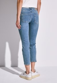 Jeans cropped blu chiaro con un design aderente, cinque tasche e vita media, abbinati a sneakers bianche su uno sfondo semplice.