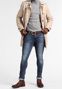 Trench coat beige con bottoni marroni, dolcevita grigio, jeans blu e stivaletti in pelle marrone. Il taglio è sartoriale e slim.
