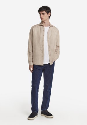 Lys beige, langærmet, knap-op skjorte med lodrette striber, båret over en hvid t-shirt. Parret med mørkeblå jeans og sorte sko.
