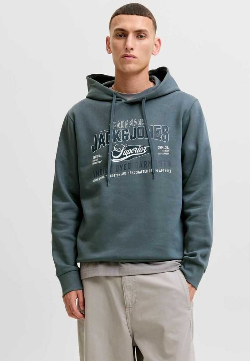 Νέος άνδρας με μπλε φούτερ Jack & Jones και μπεζ παντελόνι, στέκεται με το ένα χέρι στην τσέπη μπροστά σε απλό ανοιχτόχρωμο φόντο.