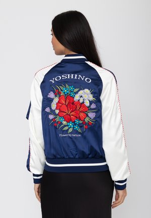 Vrouw draagt een marineblauwe en witte satijnen bomberjas met kleurrijke geborduurde bloemen en de tekst "YOSHINO Flower by nature" op de achterkant.