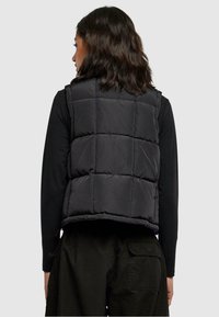 Svart puffervest, kviltet design med rektangulære mønstre, rund hals og cropped fit. Brukt over en svart langermet topp.
