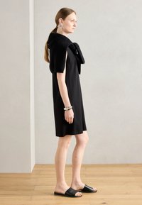 Black sleeveless dress with a textured black shawl. Silver hoop earrings and a chunky bracelet. Simple black slide sandals. Natural wooden floor.