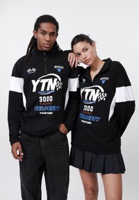 Black zip-up sweatshirts with white and blue graphics. One model wears dark jeans; another wears a pleated black skirt.