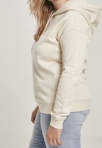 Femme portant un sweat à capuche couleur crème et un jean bleu clair, vue de profil sur un fond uni.