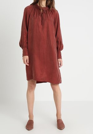Femme portant une robe ample, à manches longues, de couleur rouille, longueur genou, avec un col froncé et des chaussures plates assorties sur un fond uni.
