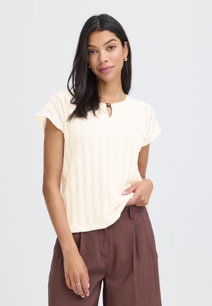 Frau mit langen dunklen Haaren, die ein beigefarbenes, strukturiertes kurzärmeliges Oberteil mit Knopfdetail und hoch taillierte braune Hose trägt, stehend vor einem einfachen Hintergrund.