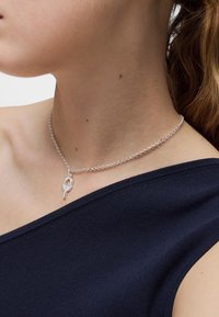 Collar de cadena de plata con un delicado colgante en forma de nudo. La cadena tiene un diseño simple con eslabones cilíndricos; el colgante presenta un diseño entrelazado.
