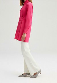 Vestido camisero fucsia de manga larga con cintura ceñida y detalles de botones, combinado con pantalones acampanados blancos y zapatos de tacón en gris claro.