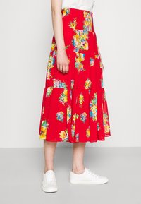 Blumen-Midikleid in leuchtendem Rot mit gestuften Design. Verfügt über bunte Blumenmuster. Getragen mit weißen Sneakers, was einen lässigen Look zeigt.