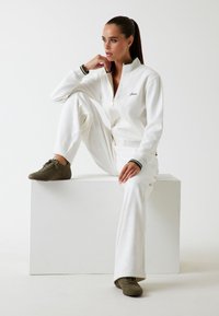 Witte jumpsuit met ritssluiting en ribbelstructuur, voorzien van tonale accenten. Groengestreepte manchetten en olijfgroene schoenen. Model zittend op een witte sokkel.