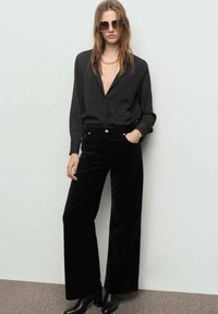 Pantalones de terciopelo negro de pierna ancha combinados con una blusa negra de manga larga, con un escote en V profundo; complementados con gafas de sol extragrandes.