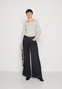 Graues Langarmoberteil kombiniert mit dunkelblauen weit geschnittenen Jeans. Die Jeans haben eine hohe Taille und auffällige Nähte. Schwarze Stiefel und eine kleine Tasche sind ebenfalls enthalten.
