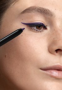 Crayon eye-liner violet près de l'œil, avec une application audacieuse en aile d'oiseau. Gros plan sur la peau avec de légères taches de rousseur, une texture lisse et une couleur naturelle des lèvres.