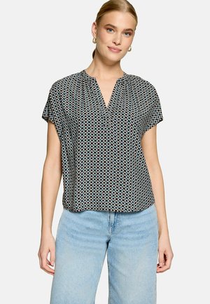 Femme portant un chemisier à manches courtes à motifs avec un col en V et un jean bleu clair, debout devant un fond uni.