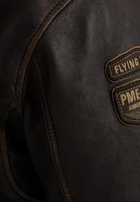 Brun læderjakke med et nærbillede af tekstureret materiale, kontrastsyninger og to stofmærker med prægede bogstaver.