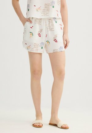 Mujer con pantalones cortos crema con estampados temáticos de comida y top sin mangas a juego, combinados con sandalias deslizantes de tejido beige, de pie con las manos en los bolsillos.