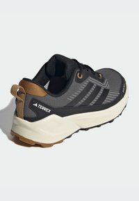 Chaussure de sport noire et grise avec une tige texturée, trois rayures noires, un accent beige au niveau du talon et une semelle extérieure légère couleur crème.