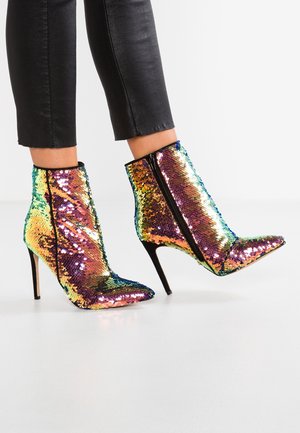 Stopy w czarnych spodniach noszące botki za kostkę z ostro zakończonym noskiem, pokryte błyszczącymi, wielokolorowymi cekinami, na szpilkach i z czarnymi zamkami błyskawicznymi.