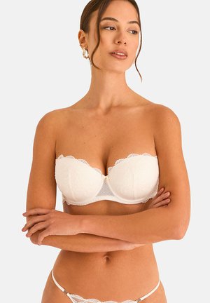 Femme portant un soutien-gorge en dentelle blanc sans bretelles et un string en dentelle assorti, les bras croisés, regardant légèrement sur le côté sur un fond uni.