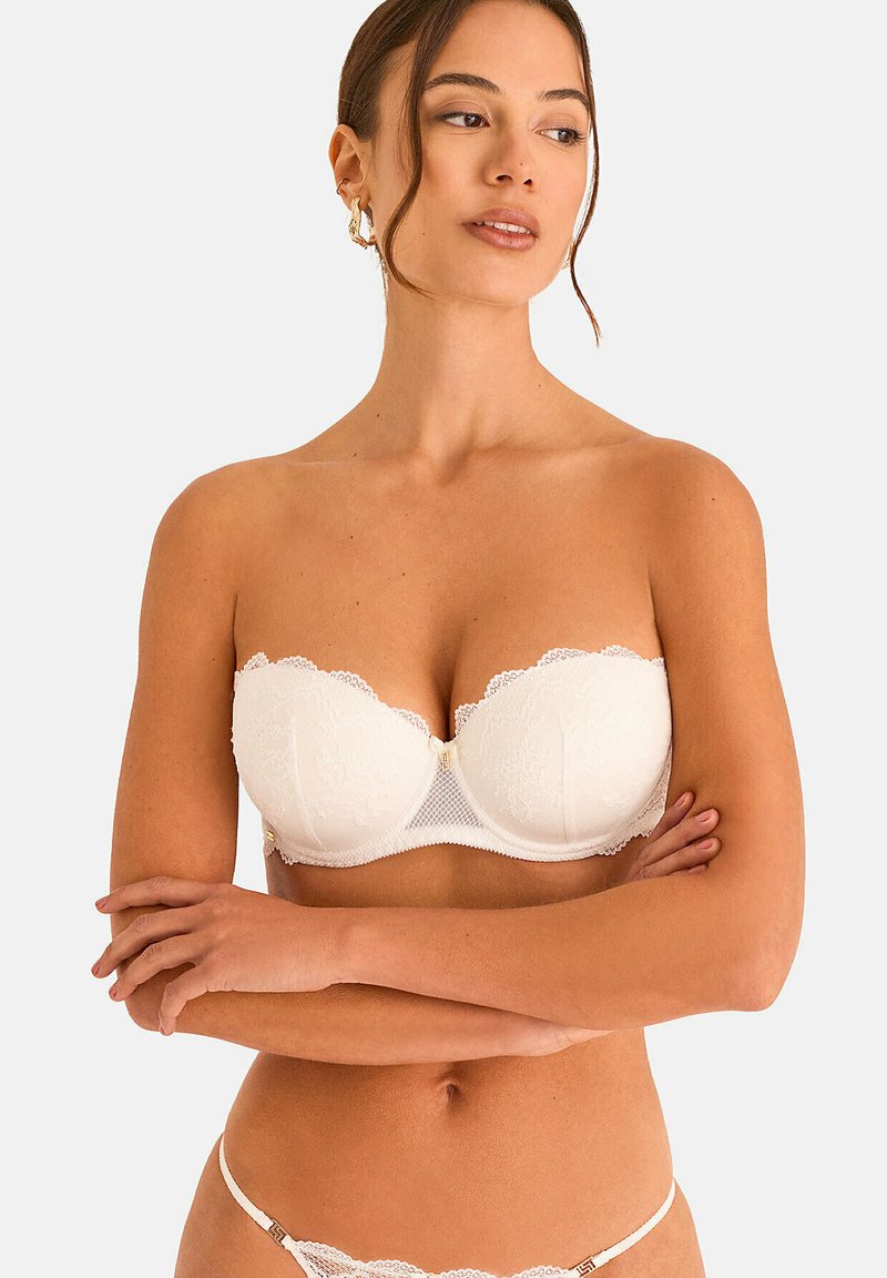Femme portant un soutien-gorge en dentelle blanc sans bretelles et un string en dentelle assorti, les bras croisés, regardant légèrement sur le côté sur un fond uni.