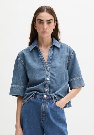 Femme aux longs cheveux bruns portant une chemise en jean ample à manches courtes et un jean bleu taille haute, une main dans la poche.