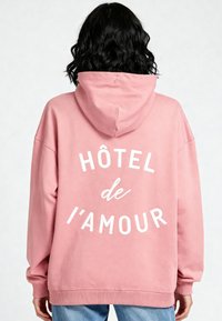 Felpa rosa con una grande grafica testuale bianca "HÔTEL de l'amour" sul retro. Presenta un cappuccio e polsini a costine, realizzata in un tessuto morbido.