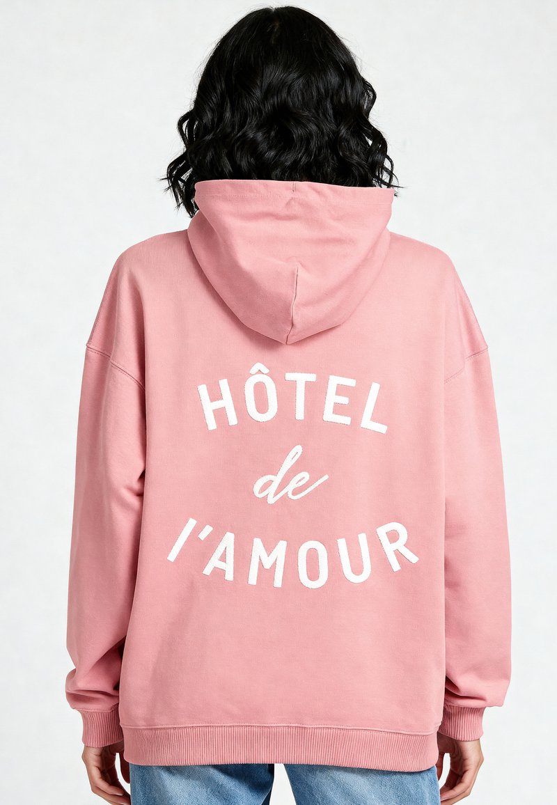 Felpa rosa con una grande grafica testuale bianca "HÔTEL de l'amour" sul retro. Presenta un cappuccio e polsini a costine, realizzata in un tessuto morbido.