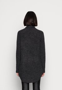 Person mit kurzem, dunklem Haar, der einen langen, dunkelgrauen, gestrickten Pullover und schwarze Hosen trägt und vor einem schlichten, hellgrauen Hintergrund steht.