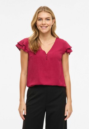 Naine naeratab, kannab lühikeste varrukatega tumeroosat pluusi, millel on nööpide detail, ja musta pükse, seisab valge tausta ees.