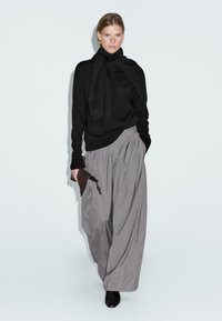 Maglione nero a maniche lunghe con dettaglio a sciarpa, abbinato a pantaloni grigi a pieghe ampi. La modella tiene una piccola pochette marrone, indossando scarpe nere.