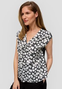 Zwart-witte blouse met korte mouwen en v-hals, met een textuur van stippen en een knoopdetail aan de voorkant. De gladde stof valt soepel.