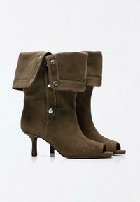 Braune Wildleder-Stiefel mit mittelhohem Absatz bis zur Mitte der Wade, umgeschlagenen Manschetten, silbernen Druckknöpfen und offenem Zeh-Design auf weißem Hintergrund.