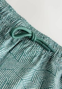 Detail elastických šortiek so zeleno-bielym vzorom na pásovej časti, s uviazaným zeleným šnúrkou, na koncoch ktorej je nápis „NEXT“.