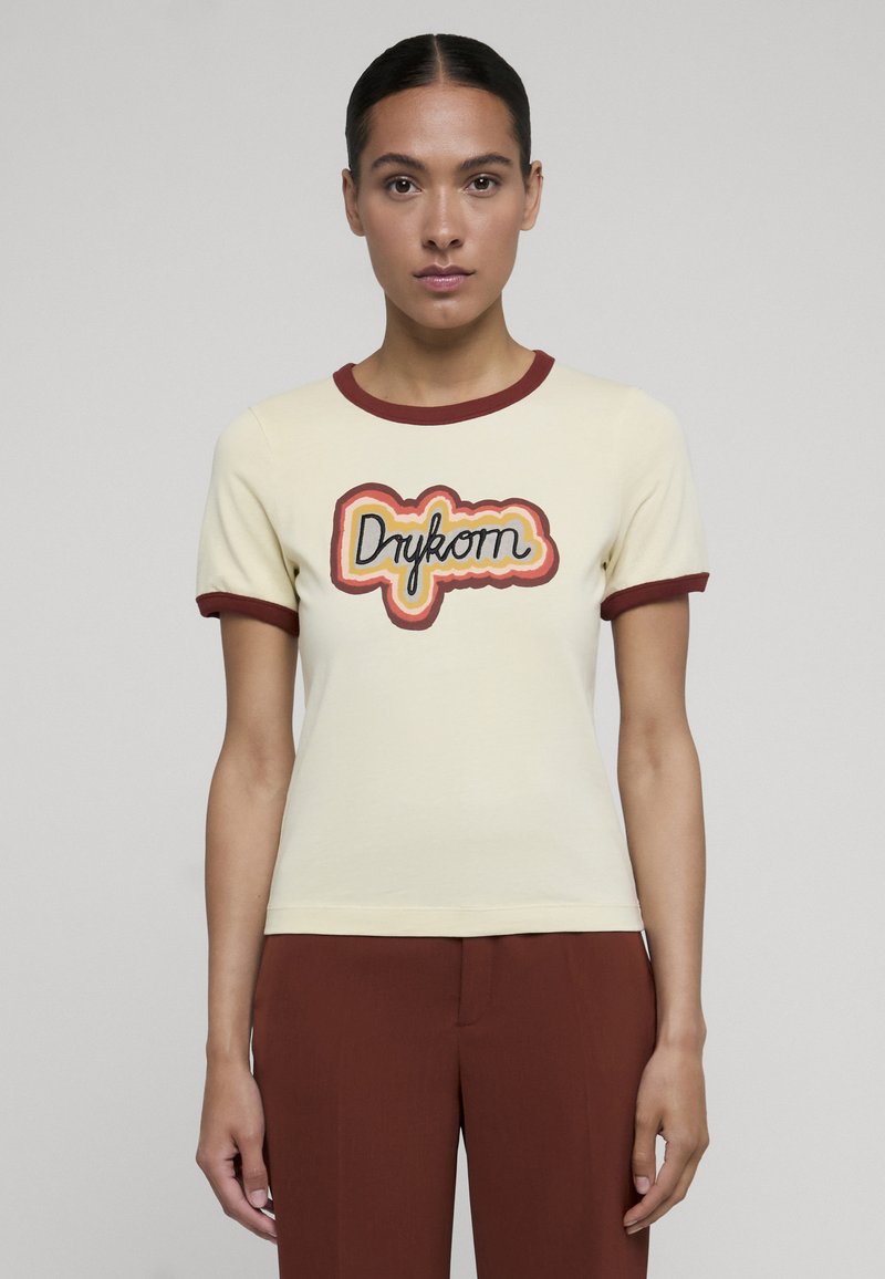 Femme debout face à l'avant, portant un chemisier crème à manches courtes avec des bordures bordeaux et le logo "Drykorn", assorti à un pantalon bordeaux.