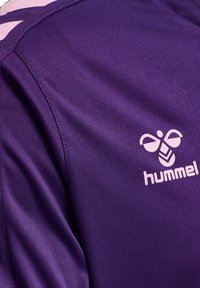 Lilla sporttrøje med en glat tekstur, korte ærmer og et pink "hummel" logo på den øverste del af ryggen.