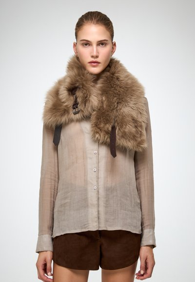Donna che indossa una camicia trasparente beige con bottoni, pantaloni corti marroni e un grande collare di pelliccia marrone soffice con cinturini in pelle, su uno sfondo neutro.