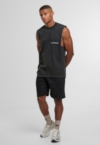 Maglietta senza maniche nera con scollo rotondo e logo bianco, abbinata a pantaloni corti neri. Il modello indossa scarpe da ginnastica chiare e calzini beige.