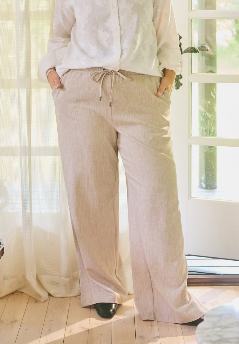Personne portant un pantalon beige à jambes larges avec un cordon de serrage et une chemise blanche à manches longues, debout à l'intérieur près d'une porte les mains dans les poches.