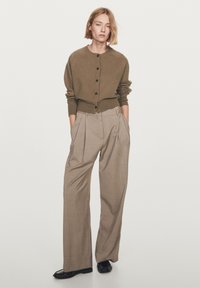 Brun stickad kofta med knappar, kombinerad med ljusbeige plisserade byxor. Outfitten har en avslappnad passform och mjuka texturer. Svarta skor kompletterar looken.